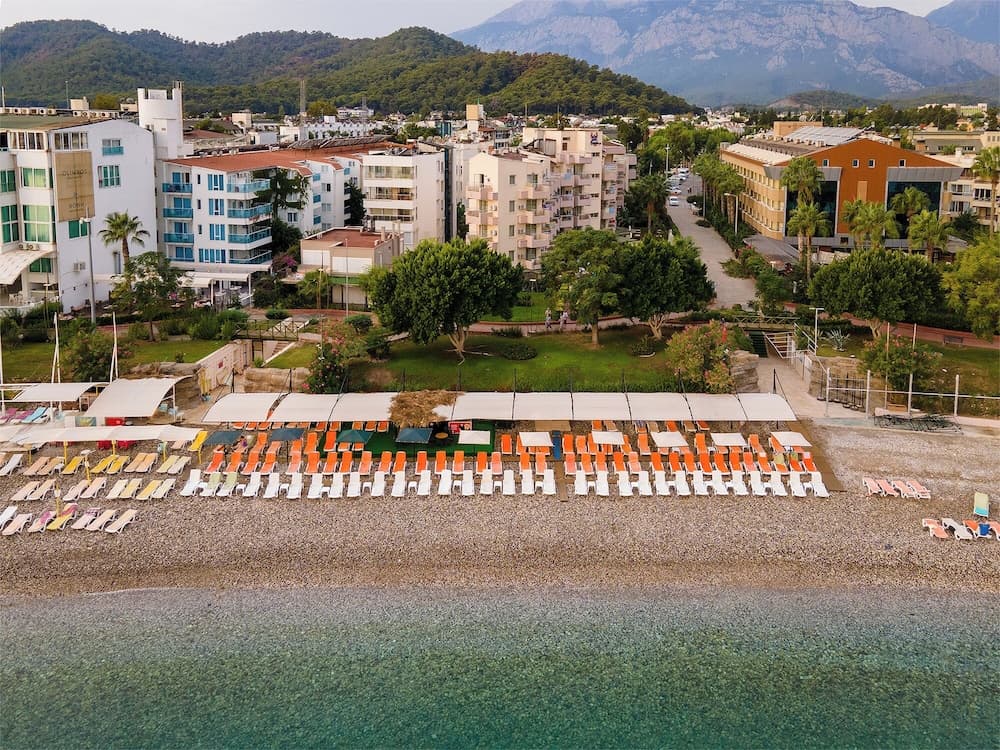 A serene coastal resort scene features colorful lounge chairs on a pebble beach, surrounded by lush greenery and modern buildings, with majestic mountains in the background, ideal for travelers seeking relaxation and scenic beauty.