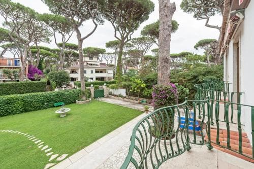 A serene balcony overlooks a lush garden with manicured lawns, vibrant flowers, and towering pine trees, ideal for a tranquil vacation retreat.