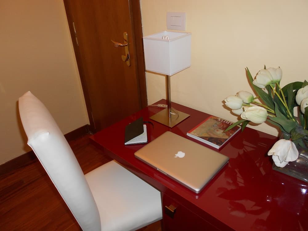 A cozy hotel room workspace features a sleek red desk adorned with a laptop, tulips, and art books, complemented by a white chair and a modern lamp, ideal for travelers seeking productivity and comfort.