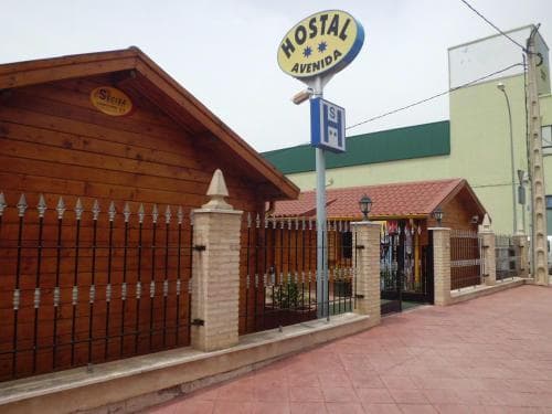 A cozy hostel entrance with a wooden facade and metal gate invites travelers in a quiet urban area, featuring a bright "Hostal Avenida" sign and a welcoming atmosphere ideal for budget-friendly stays.