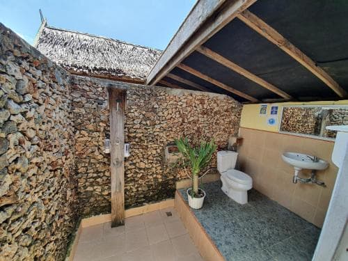 An outdoor bathroom with a rustic stone wall, thatched roof, and tropical plant creates a serene, eco-friendly retreat ideal for travelers seeking unique accommodations in natural settings.