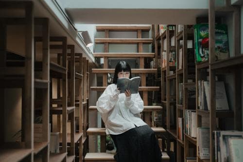 A serene library scene captures a person engrossed in reading amidst tall wooden bookshelves, ideal for travelers seeking quiet study or literary exploration.
