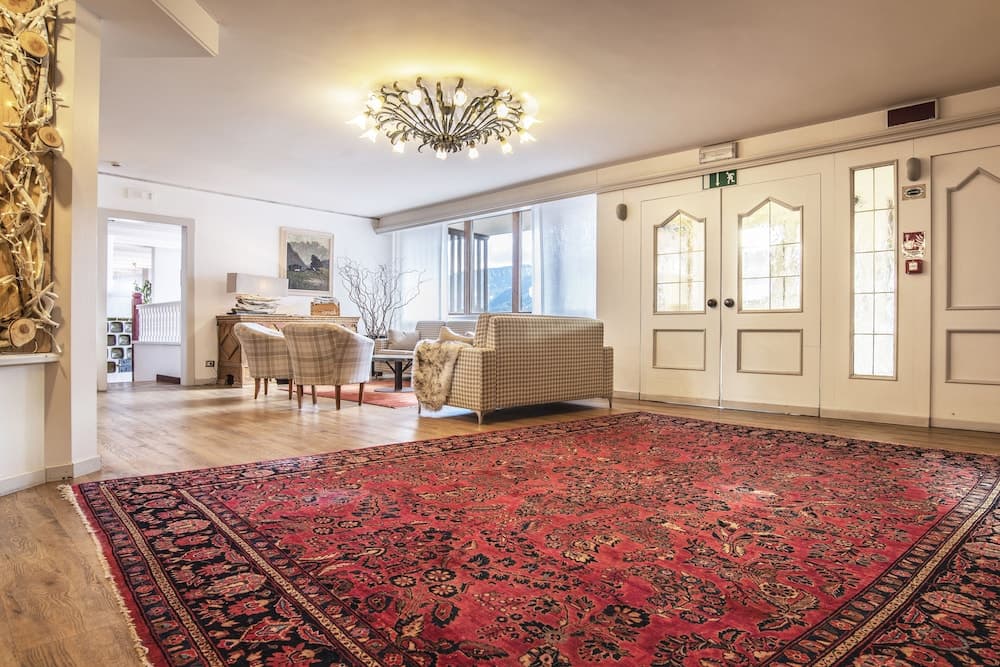 A cozy hotel lobby features a vibrant red patterned rug, elegant checkered armchairs, a sparkling chandelier, and large windows letting in natural light, ideal for travelers seeking a warm and inviting atmosphere.
