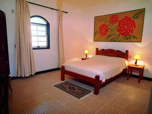 A cozy bedroom with tiled floors, a wooden bed, vibrant floral artwork, and warm lighting creates an inviting atmosphere ideal for travelers seeking a comfortable and artistic retreat.