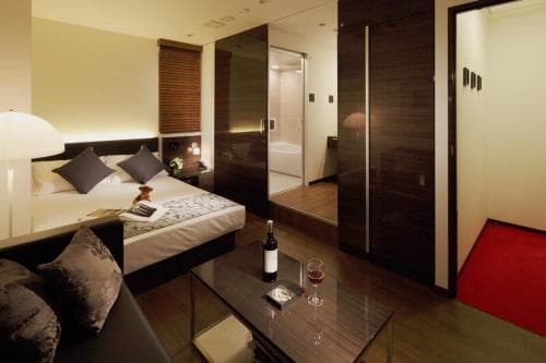 A cozy hotel suite features a modern bedroom with a neatly made bed, a glass table with wine, and an open view into a bathroom, creating a luxurious and inviting atmosphere ideal for travelers seeking comfort and relaxation.