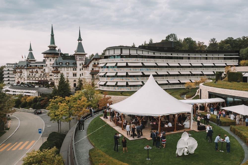 A grand hotel complex with a festive white tent set up on lush green lawns, surrounded by autumnal trees and modern architecture, creates an inviting atmosphere perfect for travelers seeking luxury events or conferences.