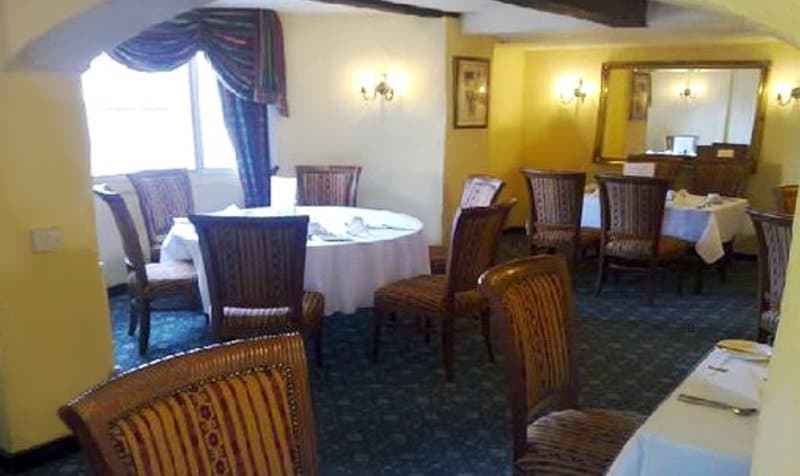 A cozy restaurant interior features patterned chairs around round tables draped in white linens, set against warm yellow walls with a large mirror and window curtains, ideal for travelers seeking a comfortable dining experience.