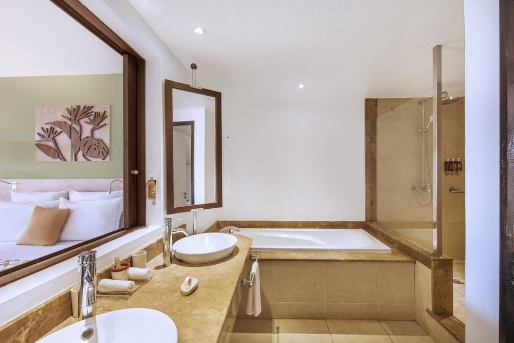 A serene hotel bathroom features a luxurious bathtub, modern sink, glass shower enclosure, and a cozy bedroom view through an open doorway, ideal for travelers seeking comfort and relaxation.