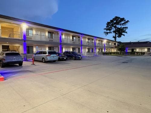 A tranquil evening at a motel with purple-lit pillars, parked cars, and a lone tree in the background, ideal for travelers seeking a cozy overnight stay.