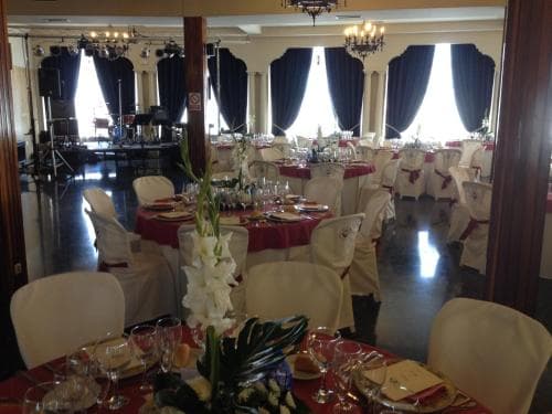 An elegant banquet hall set up for a formal event features tables adorned with red tablecloths, white chairs, floral centerpieces, and chandeliers, ideal for wedding receptions or corporate gatherings.