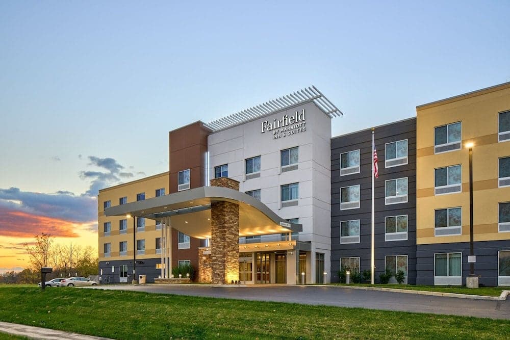A modern Fairfield by Marriott Inn & Suites hotel stands invitingly at dusk, featuring a well-lit entrance, American flag, and colorful facade, ideal for travelers seeking comfortable accommodations in a serene suburban setting.