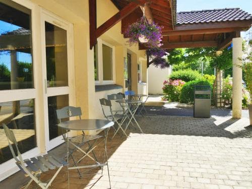A cozy outdoor patio with metal tables and chairs under a wooden awning, surrounded by lush greenery and vibrant flowers, offers a serene spot for travelers to relax and enjoy the sunny ambiance of a quaint countryside retreat.