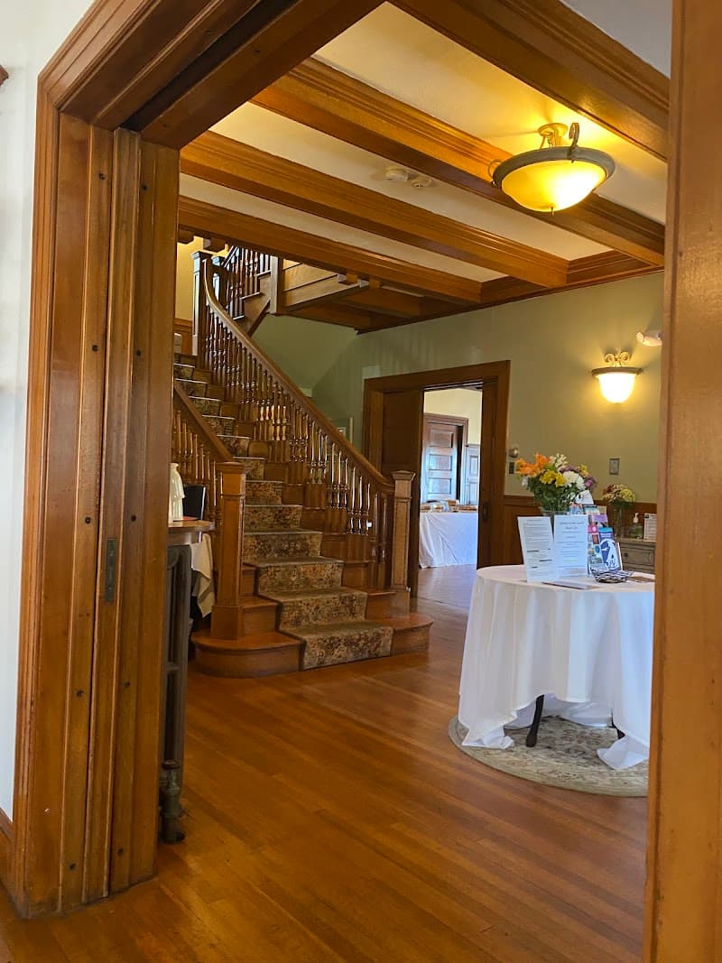 A warmly lit interior of a historic inn features polished wooden floors, a grand staircase, and a cozy reception area with floral arrangements, ideal for travelers seeking a charming and inviting accommodation experience.