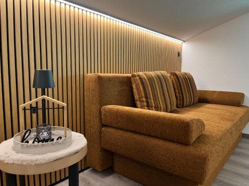 A cozy modern lounge features a plush mustard-colored sofa against a warmly lit wooden slat wall, complemented by a stylish side table with decorative items, ideal for travelers seeking a chic and relaxing accommodation space.
