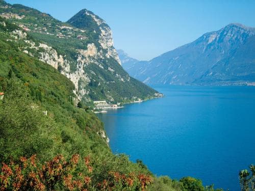 A serene lakeside vista with lush green cliffs and distant mountains under a clear blue sky, ideal for tranquil hiking and scenic photography.