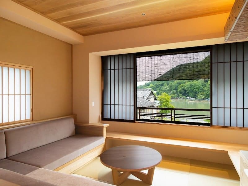 A serene Japanese ryokan room with traditional sliding doors and tatami seating offers a tranquil view of lush greenery and a distant temple, ideal for travelers seeking cultural immersion and relaxation.