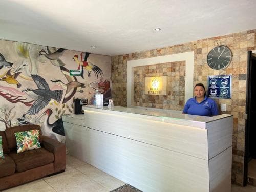 A welcoming hotel lobby features a vibrant bird mural, a modern reception desk with a friendly staff member, a cozy sofa with tropical pillows, and a stone wall with a clock, ideal for travelers seeking comfort and local charm.