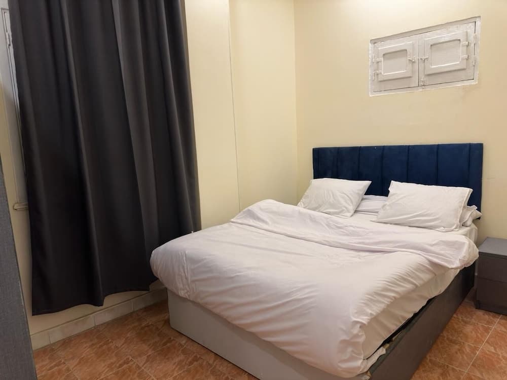 A cozy hotel room features a neatly made bed with white linens against a navy blue headboard, set beside a dark curtain and tiled floor, ideal for travelers seeking comfort and simplicity.