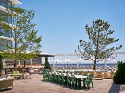 A serene seaside deck features rows of green chairs and tables under clear skies, flanked by tall trees and modern buildings, ideal for travelers seeking a relaxing coastal retreat.