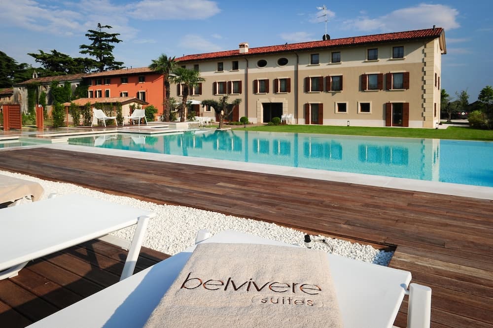 A serene resort scene features a sparkling pool surrounded by wooden decking and lounge chairs, set against the backdrop of a charming building with terracotta roofs, ideal for a relaxing vacation getaway.