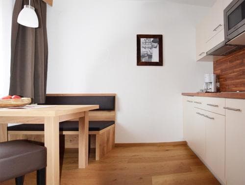 A cozy apartment kitchenette features a wooden dining table with cushioned benches, sleek white cabinetry, and warm lighting, ideal for travelers seeking a comfortable self-catering stay.