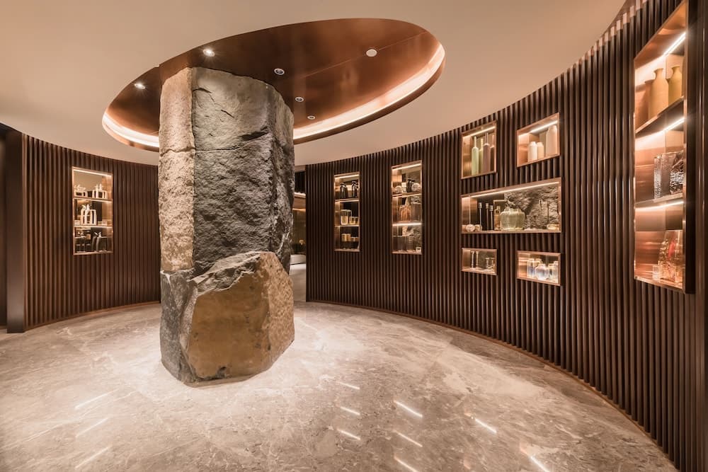A modern, luxurious interior space features a striking central rock formation under a copper-toned ceiling, surrounded by sleek wooden walls with illuminated shelves displaying elegant glassware, ideal for high-end hospitality or boutique retail environments.
