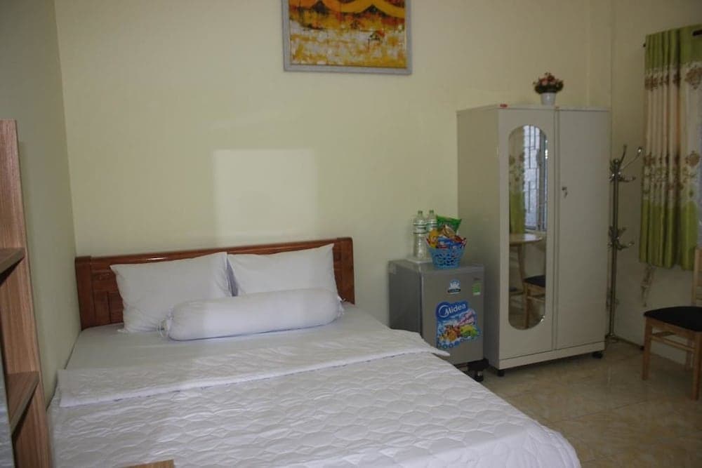 A cozy hotel room features a neatly made bed, a small fridge stocked with snacks, a mirrored wardrobe, and a vibrant wall painting, ideal for travelers seeking comfort and convenience.
