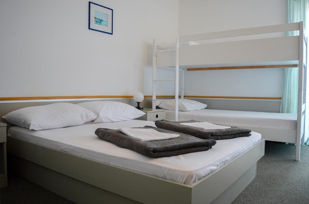 A cozy hostel room features a neatly made double bed with folded towels, a bunk bed, soft lighting, and a framed picture on the wall, ideal for budget travelers seeking comfort and simplicity.