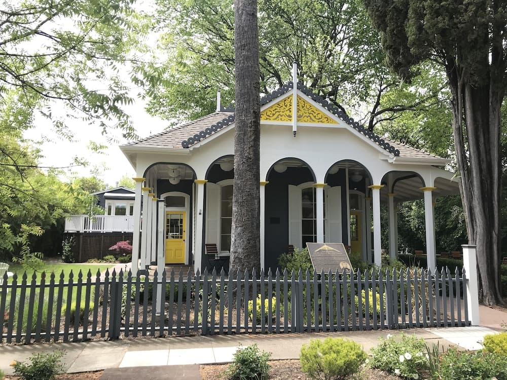 A serene park setting features a charming white pavilion with yellow accents, surrounded by lush greenery and a picket fence, ideal for leisurely strolls and cultural exploration.