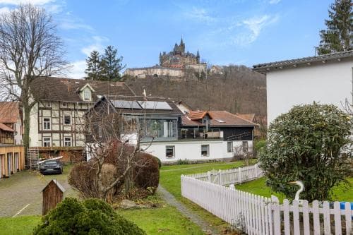 A quaint village scene with half-timbered houses and a white picket fence leads the eye to an imposing castle perched atop a hill in the distance, ideal for travelers seeking a picturesque European retreat.