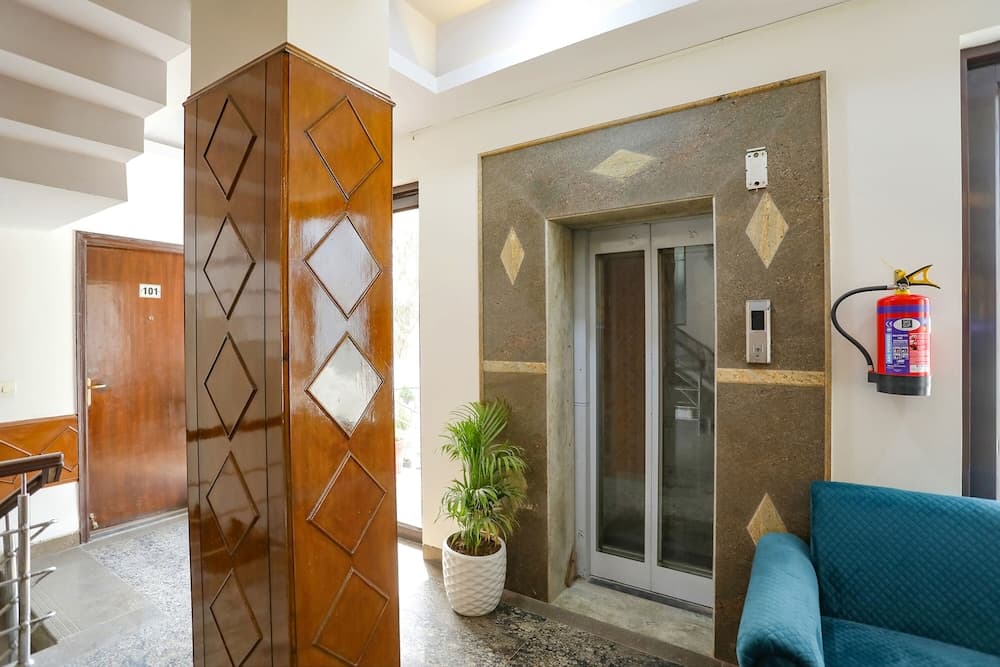A cozy hotel lobby features a modern elevator framed by marble walls, a vibrant blue sofa, a potted plant, and a fire extinguisher, ideal for travelers seeking comfort and safety during their stay.