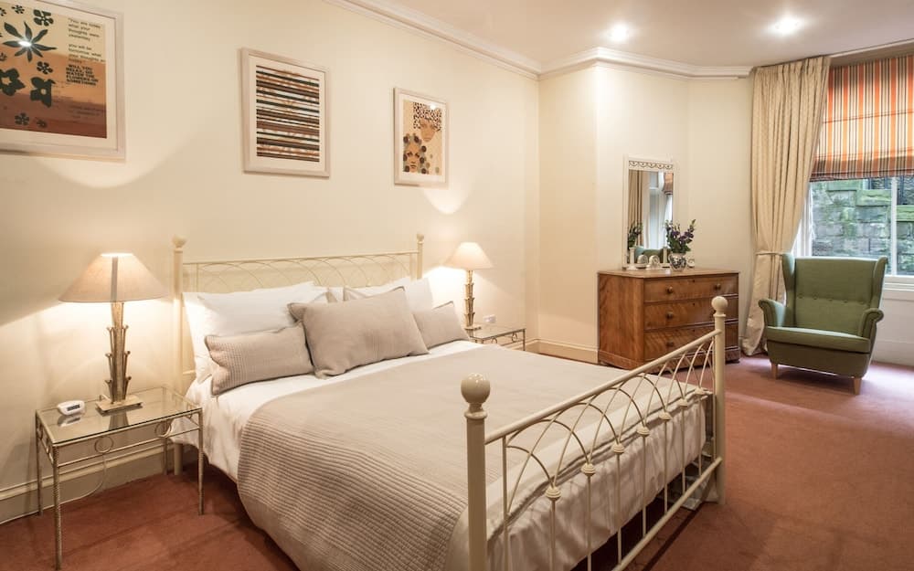 A cozy hotel bedroom features a white metal bed adorned with plush pillows, framed artwork on cream walls, a green armchair by the window, and warm lighting, ideal for travelers seeking comfort and relaxation.
