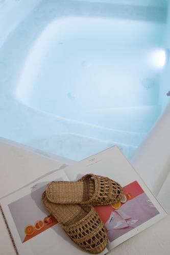 A serene spa setting features a modern bathtub filled with clear water, complemented by woven sandals resting on a colorful magazine, evoking relaxation and luxury for wellness retreats.