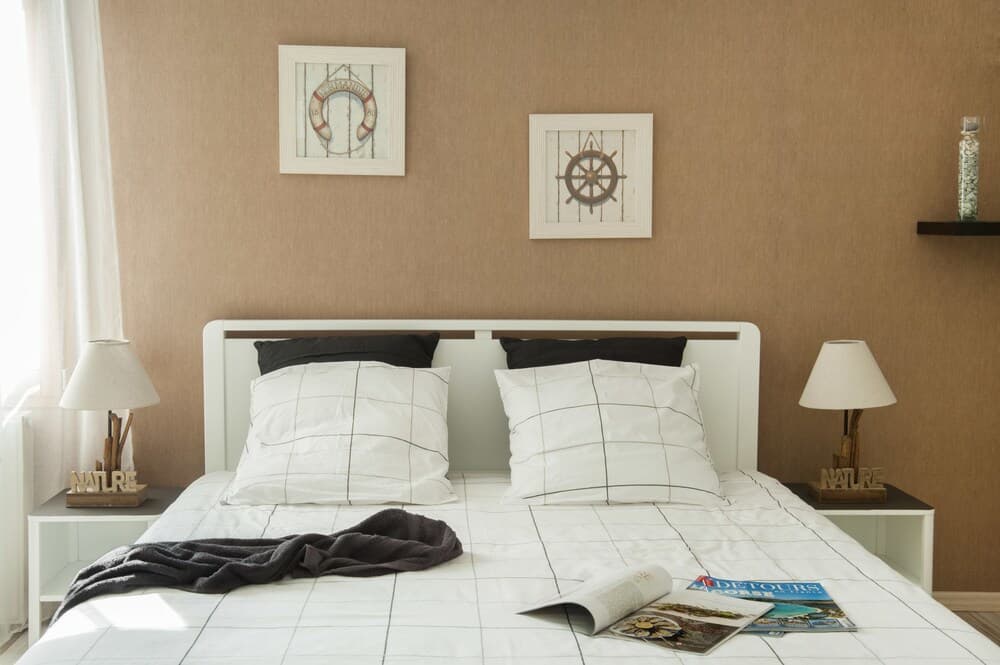 A cozy bedroom with nautical-themed decor, featuring framed lifebuoy and ship's wheel art above a neatly made bed adorned with magazines and a blanket, ideal for travelers seeking a serene and stylish retreat.