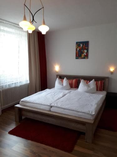 A cozy hotel room features a neatly made wooden bed with red accents, soft lighting from wall sconces, and a vibrant painting, ideal for travelers seeking comfort and relaxation.