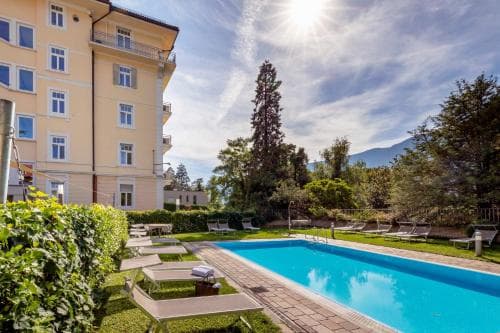 A serene hotel courtyard features a sparkling pool surrounded by lush greenery and lounge chairs under the bright sun, ideal for a relaxing vacation getaway.