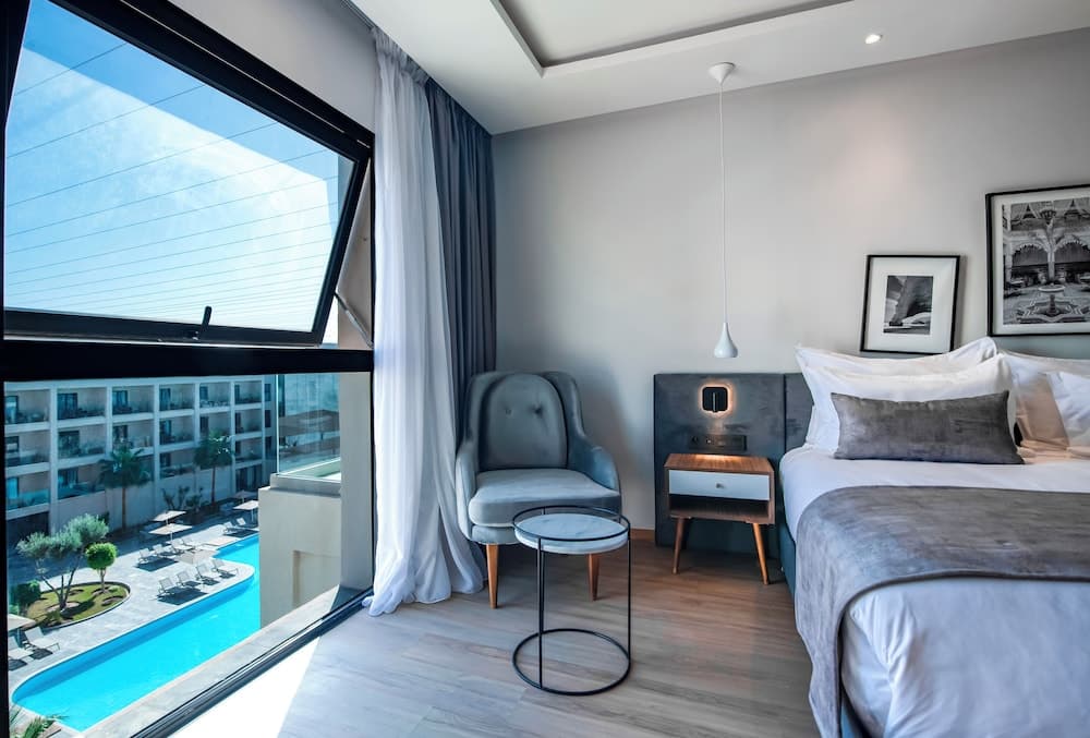 A modern hotel room with a cozy bed, stylish armchair, and large window overlooking a vibrant poolside area, perfect for travelers seeking relaxation and comfort.