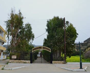 A serene entrance to a bungalow resort features lush greenery, a gated archway with "Bungalows Gouves" signage, and a tranquil suburban setting ideal for a peaceful getaway.
