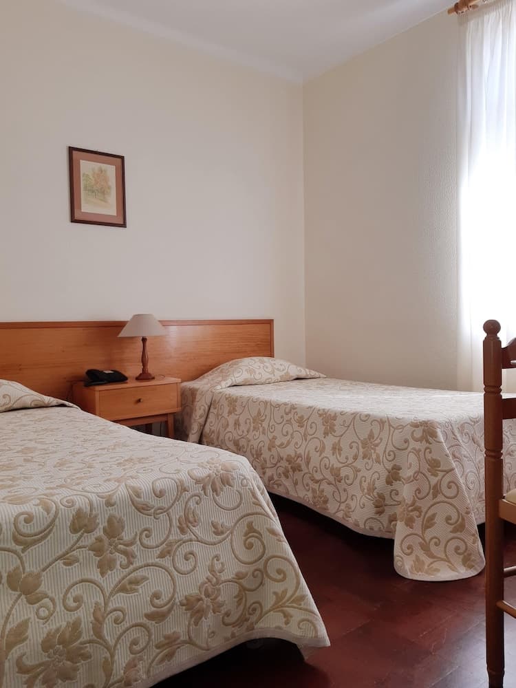 A cozy hotel room features two neatly made beds with floral-patterned bedding, a wooden nightstand with a lamp, and soft natural light filtering through sheer curtains, ideal for travelers seeking comfort and simplicity.