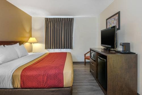 A cozy hotel room features a neatly made bed with red accents, a small desk with a TV, and warm lighting, creating an inviting atmosphere ideal for travelers seeking comfort and relaxation.