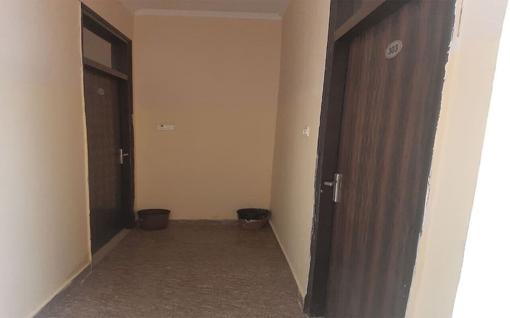 A simple hotel corridor with beige walls, wooden doors numbered "303," and two planters on the floor evokes a quiet, budget-friendly travel atmosphere ideal for solo travelers seeking basic accommodations.