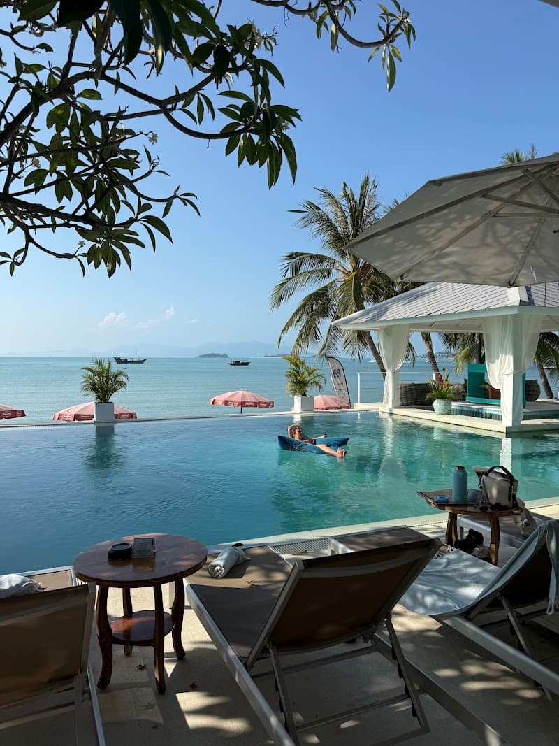 A serene tropical beach resort scene features lounge chairs, a tranquil infinity pool, and lush palm trees under a clear blue sky, ideal for relaxing vacations in Thailand.