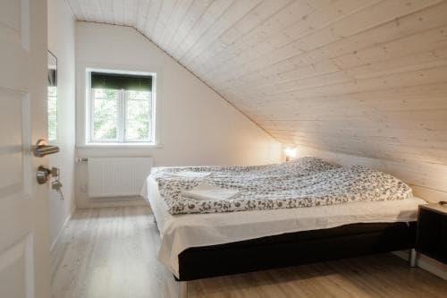 A cozy attic bedroom with wooden walls and flooring, featuring a neatly made bed by a window with greenery outside, ideal for a serene getaway or vacation rental listing.