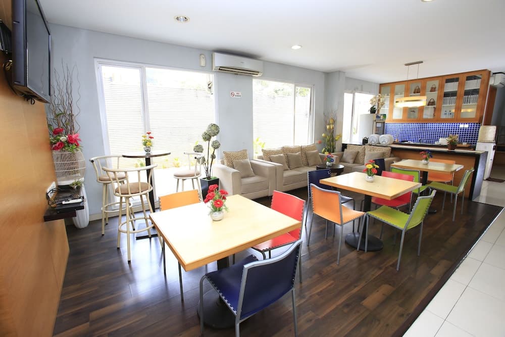 A cozy café with vibrant multi-colored chairs, wooden tables adorned with flower arrangements, large windows letting in natural light, and a kitchen area in the background, ideal for travelers seeking a relaxing spot to dine or work.