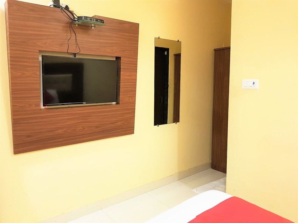 A cozy hotel room features a mounted TV on a wooden panel, a full-length mirror, and a neatly made bed with red accents, ideal for travelers seeking comfort and simplicity.