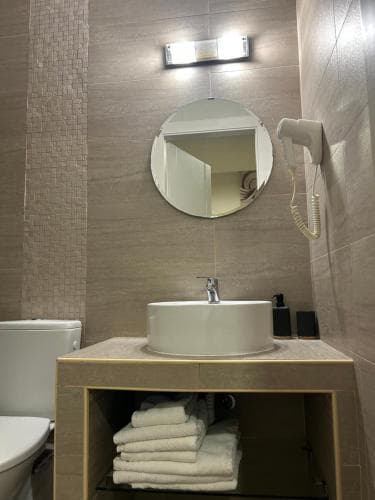 A modern hotel bathroom features a sleek sink vanity with neatly folded towels, a round mirror, and a wall-mounted hairdryer, creating a clean and inviting atmosphere ideal for travelers seeking comfort and style.
