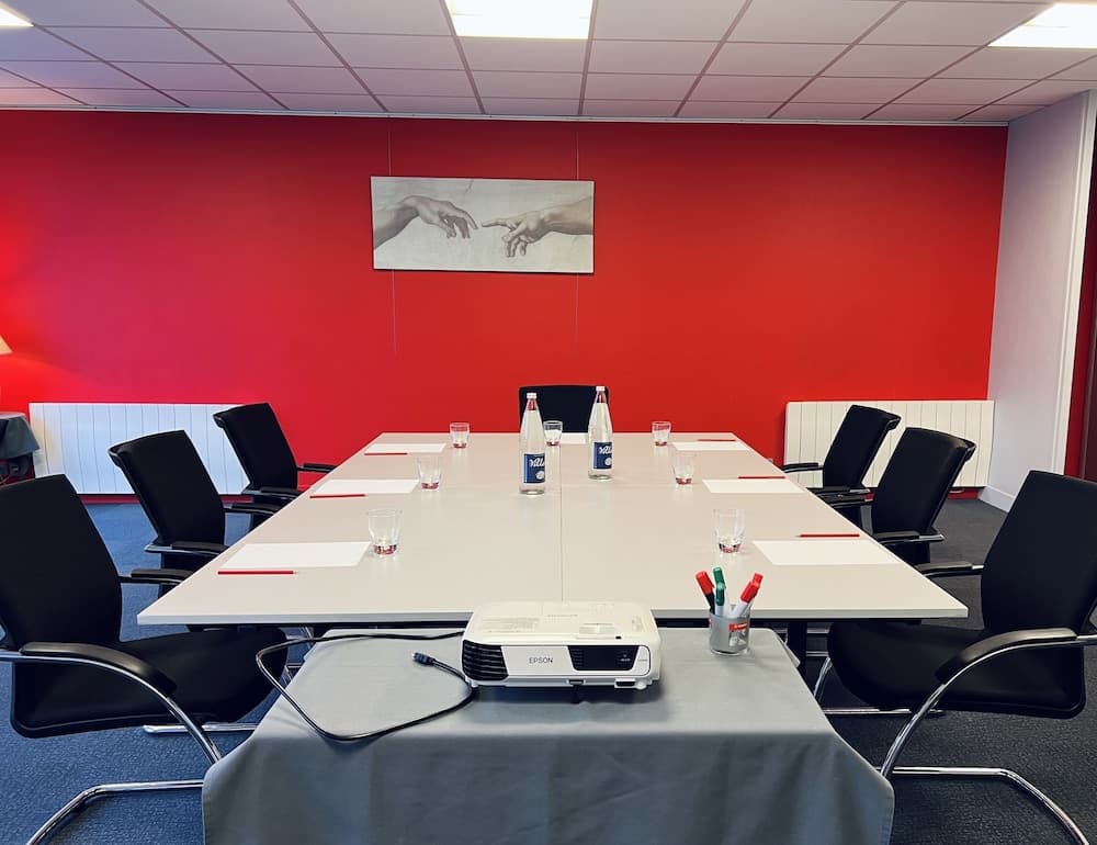 A modern conference room with a vibrant red wall, a central white table set with papers, pens, water bottles, and glasses, features an Epson projector and a striking artwork, ideal for professional meetings and presentations.