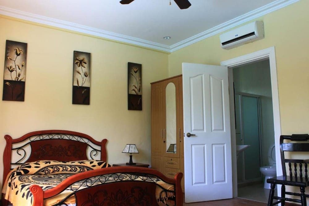 A cozy hotel bedroom with yellow walls, floral artwork, a red metal bed, and an en-suite bathroom offers travelers a comfortable and inviting retreat.