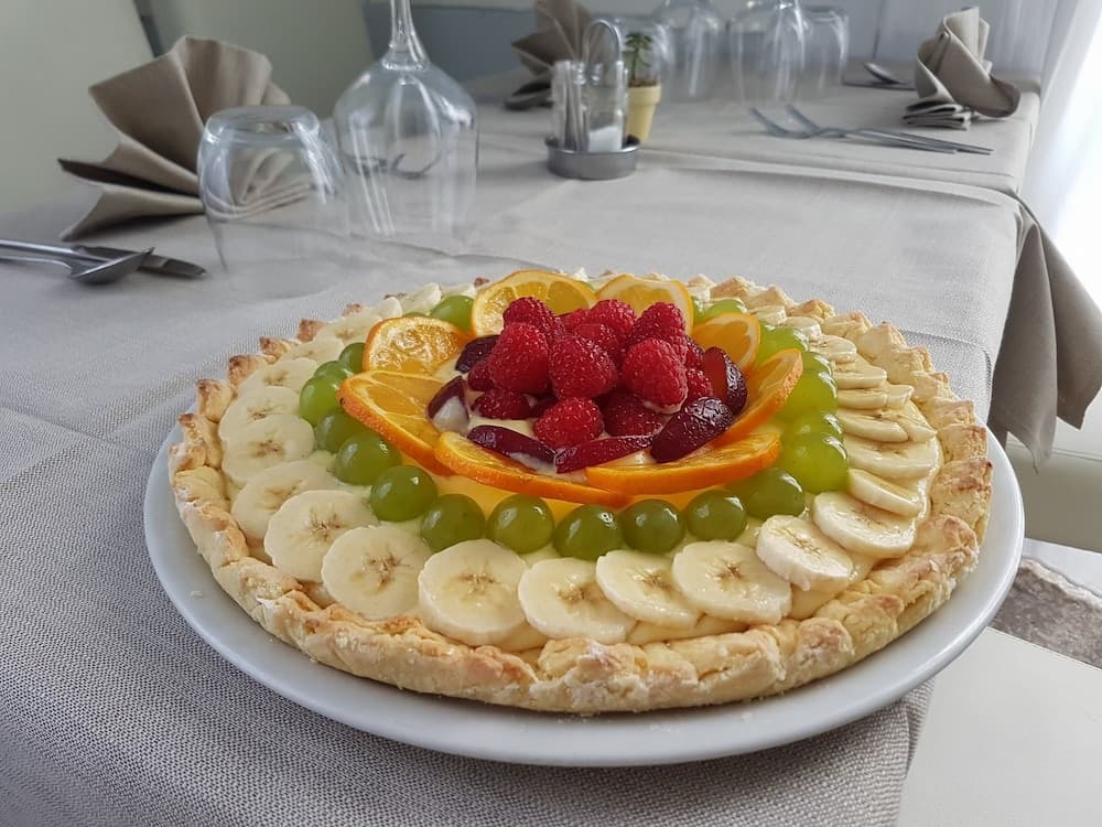 A vibrant fruit tart adorned with bananas, grapes, oranges, and raspberries sits invitingly on a neatly set dining table, perfect for a delightful culinary adventure in a cozy restaurant setting.