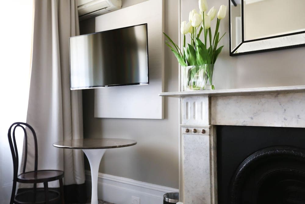 A cozy hotel room corner features a sleek fireplace adorned with white tulips, a modern flat-screen TV, and a minimalist table and chair setup, ideal for travelers seeking comfort and style.